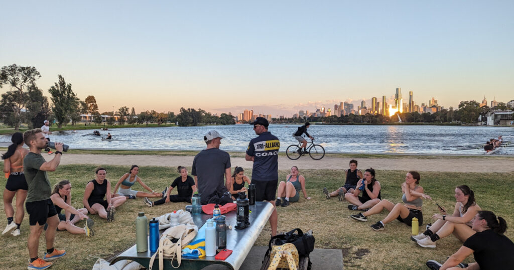 Tri Alliance coach briefing a run group at Albert Park Lake with the Melbourne skyline behind