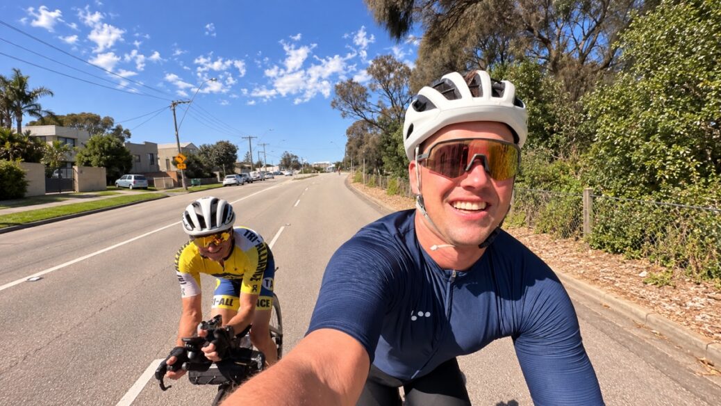 Two cyclists in triathlon attire riding on a paved road with trees and houses in the background.