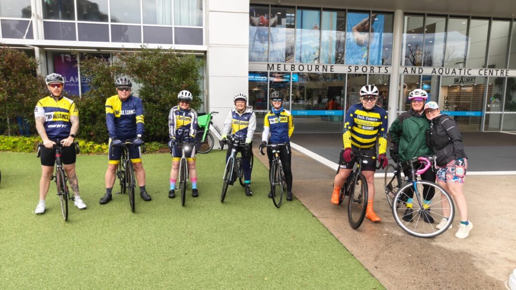 Cyclists in yellow and blue team jerseys with bicycles at Melbourne Sports and Aquatic Centre.